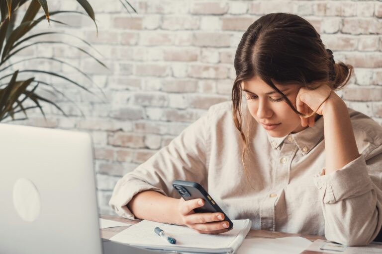 Close up millennial female student sitting at desk, holding phone in hand, checking information